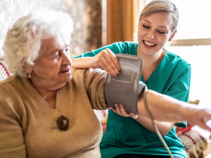 Eine junge Pflegerin legt einer älteren Frau lächelnd ein Blutdruckmessgerät an.